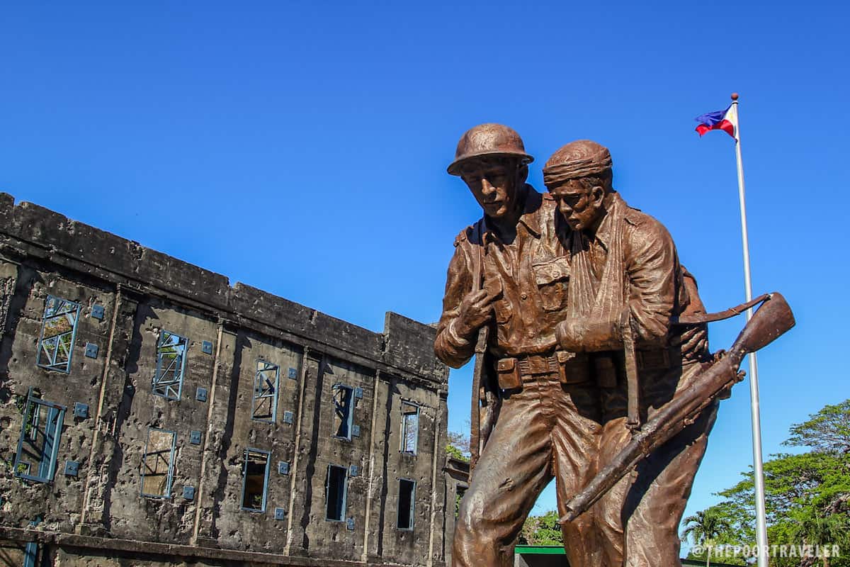 A statue of an American and a Filipino with the ruins of Cine Corregidor and the Philippine flag in the background.